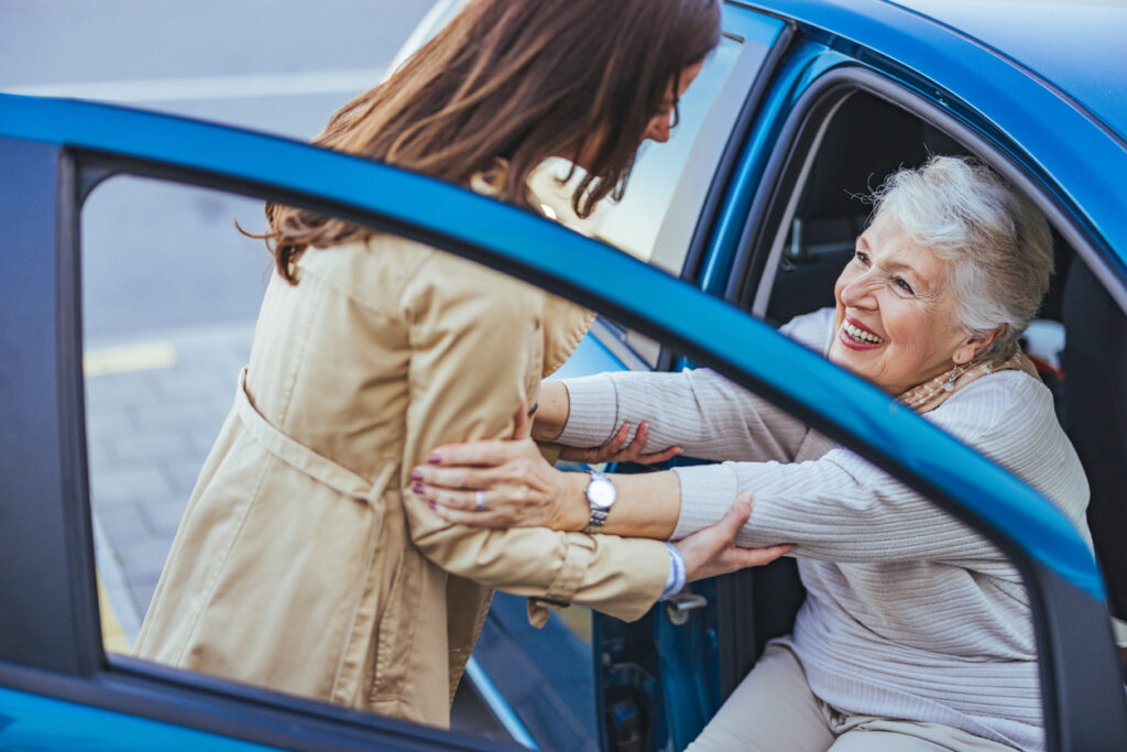 A joyful elderly woman is being helped out of a blue car by a caregiver. The scene conveys warmth, support, and care, highlighting the bond between different generations.