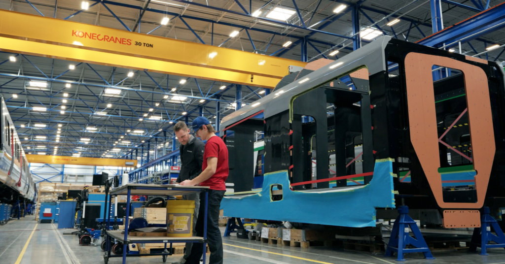 Two people working in front of a light rail vehicle being put together in a manufacturing facility. 